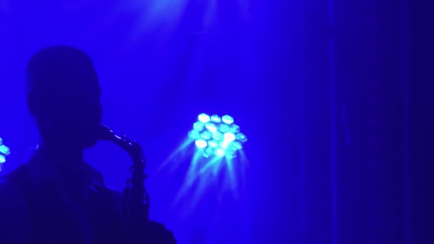 Silueta mladý retro stylový chlap hraje na zlatý lesklý saxofon v modrých reflektorech na jevišti. Přízvuk na saxofon. Tmavé studio s kouřem a neonovým osvětlením. Zavřít. — Stock video