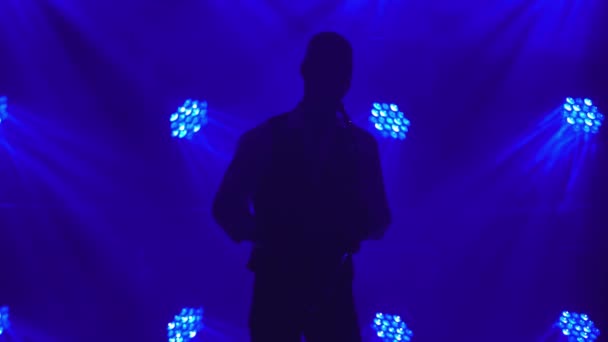 Silhouette egy fiatal retro stílusos srác játszik az arany fényes szaxofonon a kék reflektorfényben a színpadon. Sötét garzon füsttel és neon világítással. — Stock videók