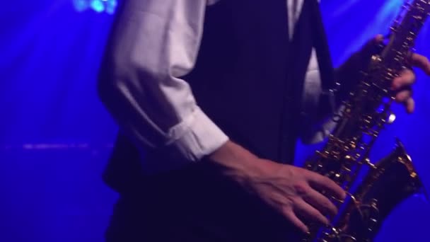 Un giovane ragazzo retrò elegante gioca sul sassofono dorato lucido sotto i riflettori blu sul palco. Studio scuro con fumo e luci al neon. Mani e sassofono da vicino. Vista laterale. — Video Stock