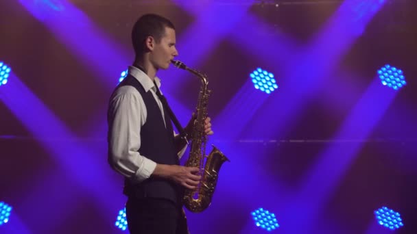 Een jonge retro stijlvolle man speelt op de gouden glanzende saxofoon in de blauwe spots op het podium. Donkere studio met rook en neon verlichting. Zijaanzicht. — Stockvideo