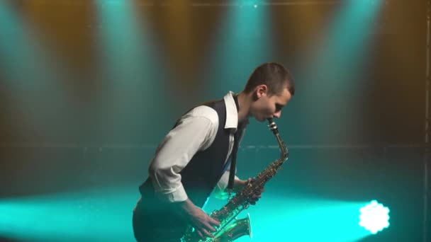 Un jeune chic rétro joue sur le saxophone doré brillant sous les projecteurs turquoise sur scène. Studio sombre avec fumée et néon. Vue latérale. — Video