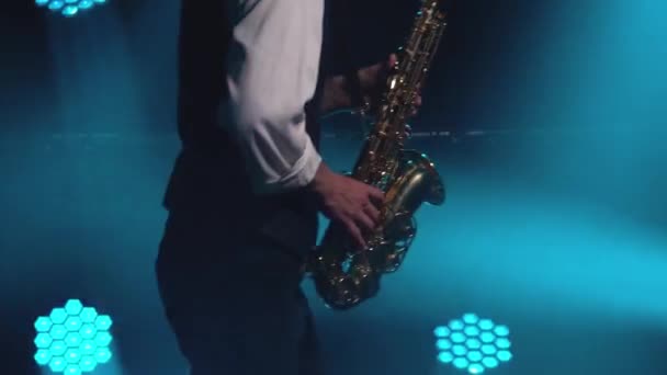 Mladý retro stylový chlap hraje na zlatý lesklý saxofon v tyrkysových reflektorech na jevišti. Tmavé studio s kouřem a neonovým osvětlením. Ruce a saxofon zblízka. Boční pohled. — Stock video