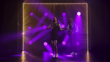 Güzel bir vokalist kız, elleri klasik bir mikrofonda sahnede şarkı söyleyip el kol hareketi yapıyor. Siyah kısa elbiseli esmer, pullu ve mor spot ışıklarının altında beyaz makosen ayakkabılı. İçeri