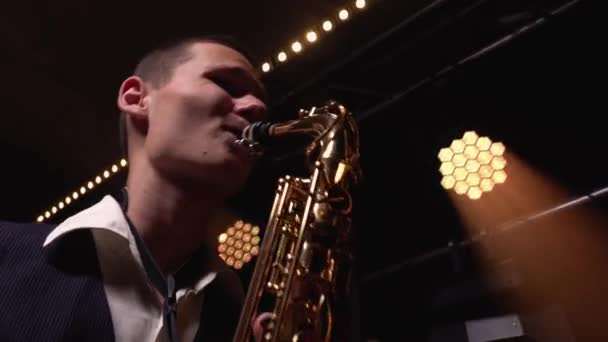 Ein junger Retro-Stylist spielt auf dem goldglänzenden Saxofon auf der Bühne. Dunkles Studio mit Rauch und Neonlicht. Ansicht von unten. Nahaufnahme. — Stockvideo