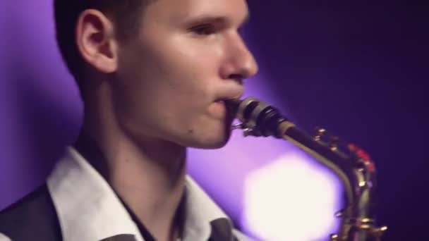 Молодий ретро-стильний хлопець грає на золотому блискучому саксофоні на сцені. Темна студія з димом і неоновим освітленням. Обличчя крупним планом. Вид збоку . — стокове відео