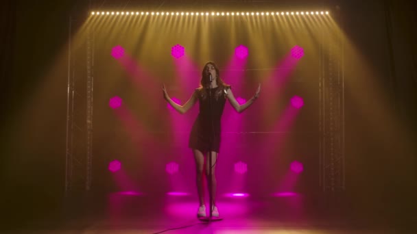 A beautiful girl vocalist singing and gestures with his hands on stage in a vintage microphone. A brunette in a short black dress with sequins and white loafers in the rays of pink spotlights. Dark — Stock Video