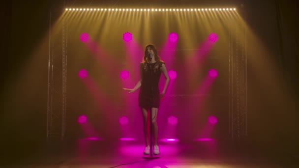Ένα όμορφο κορίτσι τραγουδίστρια και χειρονομίες με τα χέρια του στη σκηνή σε ένα vintage μικρόφωνο. Μια μελαχρινή σε ένα κοντό μαύρο φόρεμα με πούλιες και λευκά loafers στις ακτίνες του ροζ προβολείς. Σκούρο — Αρχείο Βίντεο