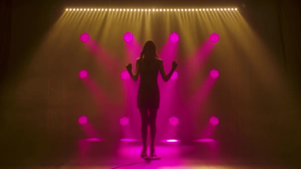 Silhouet een mooie zangeres die zingt en gebaart met zijn handen op het podium in een vintage microfoon. Een brunette in een korte zwarte jurk met pailletten en witte loafers in de stralen van roze — Stockvideo