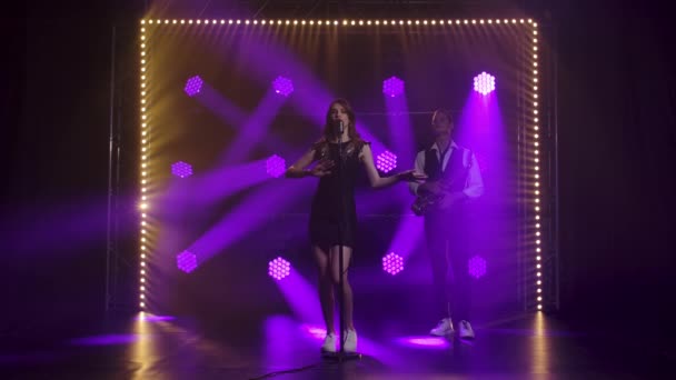 Una hermosa vocalista cantando y gestos con sus manos en el escenario en un micrófono vintage. Una morena en un vestido negro corto con lentejuelas y mocasines blancos en los rayos de focos púrpura. En — Vídeos de Stock