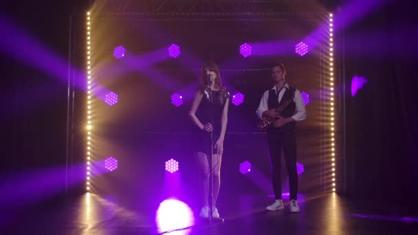 A beautiful girl vocalist singing and gestures with his hands on stage in a vintage microphone. A brunette in a short black dress with sequins and white loafers in the rays of purple spotlights. In — Stock Video