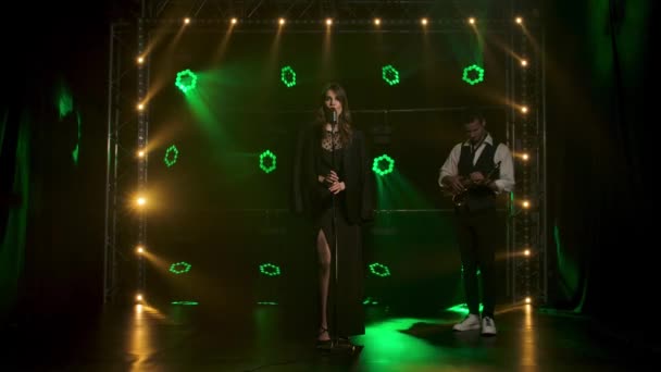 Una hermosa vocalista cantando lánguidamente y gestos con sus manos en el escenario en un micrófono vintage. Una morena con un largo vestido negro y chaqueta de hombre en los rayos de los focos verdes. En el — Vídeos de Stock