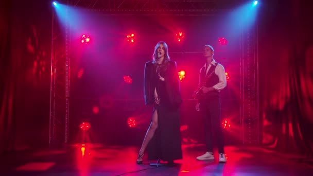 A young girl singer looks passionately at a guy saxophonist and sings a song on stage into a vintage microphone. A brunette in a black dress and a mens jacket on the shoulders with red lipstick on — Stock Video