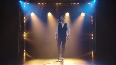 Genç stil sahibi bir vokalist elleri klasik bir mikrofonda sahnede şarkı söylüyor ve jestler yapıyor. Dumanlı ve neon ışıklı karanlık stüdyo. Dinamik neon ışık efektleri. Performans vokal ve