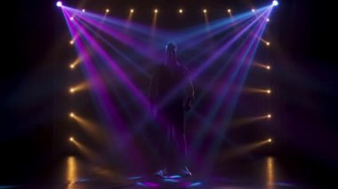 Siluet, genç stil sahibi bir vokalist, elleri klasik bir mikrofonda sahnede şarkı söylüyor ve jestler yapıyor. Dumanlı ve neon ışıklı karanlık stüdyo. Dinamik neon ışık efektleri. Performans sesi