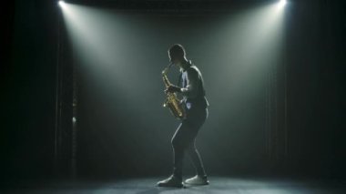 Siluet, genç ve şık bir adam sahnede altın gibi parlak bir saksafon çalıyor. Dumanlı ve neon ışıklı karanlık stüdyo. Performans vokali ve müzik grubu. Yan görüş. Yavaş çekim.