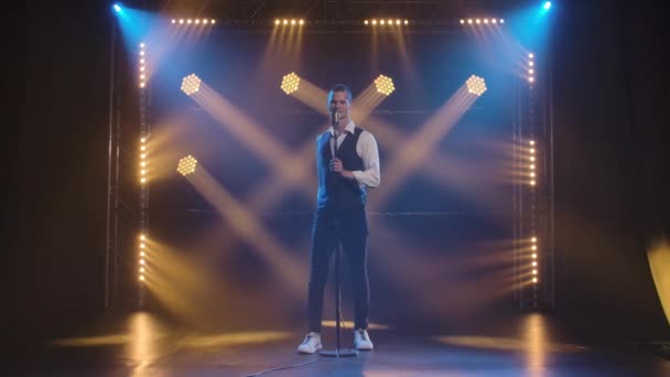 A young stylish guy vocalist singing and gestures with his hands on stage in a vintage microphone. Dark studio with smoke and neon lighting. Dynamic neon lighting effects. Performance vocal and — Stock Video