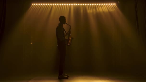 Silueta un chico joven y elegante toca el saxofón dorado brillante en los focos amarillos en el escenario. Estudio oscuro con humo e iluminación de neón. Banda vocal y musical. Vista lateral. Movimiento lento. — Vídeo de stock