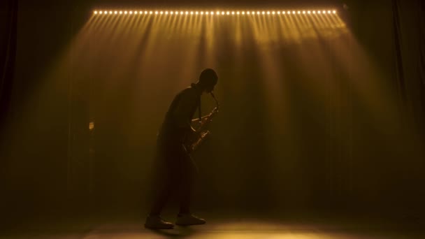 Silhouette a young stylish guy plays the golden shiny saxophone in the yellow spotlights on stage. Dark studio with smoke and neon lighting. Performance vocal and musical band. Side view. Slow motion. — Stock Video