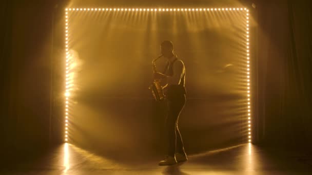 Silhouette un jeune homme élégant joue du saxophone doré brillant sous les projecteurs jaunes sur scène. Studio sombre avec fumée et néon. Performance vocale et orchestre musical. Vue latérale. Mouvement lent. — Video