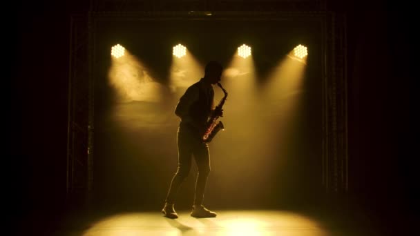 Silhouette un jeune homme élégant joue du saxophone doré brillant sous les projecteurs jaunes sur scène. Studio sombre avec fumée et néon. Performance vocale et orchestre musical. Mouvement lent. — Video