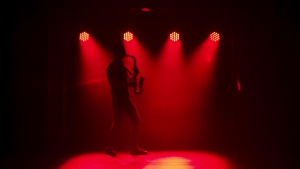 Силуэт молодой стильный парень играет золотой блестящий саксофон в разноцветных прожекторов на сцене. Темная студия с дымом и неоновым освещением. Динамические неоновые световые эффекты. Вид сбоку. Медленное движение — стоковое видео