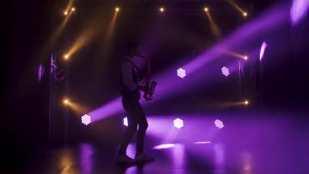 Silueta un chico joven y elegante toca el saxofón brillante dorado en los focos púrpura en el escenario. Estudio oscuro con humo e iluminación de neón. Efectos dinámicos de iluminación de neón. Vista lateral. Movimiento lento. — Vídeo de stock