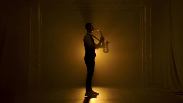 Silhouet een jonge stijlvolle man speelt de gouden glanzende saxofoon in de gele spots op het podium. Donkere studio met rook en neon verlichting. Optreden vocale en muzikale band. Zijaanzicht. Langzame beweging. — Stockvideo