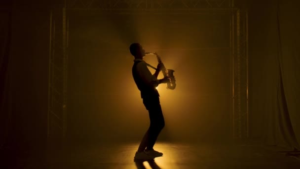 Silhouette un jeune homme élégant joue du saxophone doré brillant sous les projecteurs jaunes sur scène. Studio sombre avec fumée et néon. Performance vocale et orchestre musical. Vue latérale. Mouvement lent. — Video
