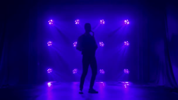 Silhouette un jeune homme élégant joue du saxophone doré brillant sous les projecteurs bleus sur scène. Studio sombre avec fumée et néon. Performance vocale et orchestre musical. Vue latérale. Mouvement lent. — Video