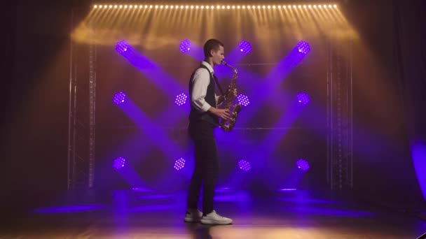 Een jonge stijlvolle man speelt de gouden glanzende saxofoon in de blauwe spots op het podium. Donkere studio met rook en neon verlichting. Neon lichteffecten. Zijaanzicht. Langzame beweging. — Stockvideo