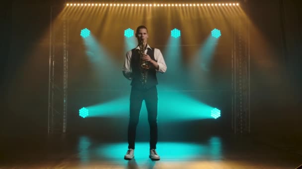 A young stylish guy plays the golden shiny saxophone in the turquoise spotlights on stage. Dark studio with smoke and neon lighting. Neon lighting effects. Slow motion. — Stock Video