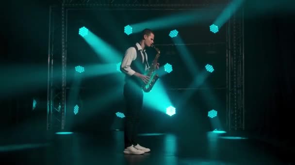 A young stylish guy plays the golden shiny saxophone in the turquoise spotlights on stage. Dark studio with smoke and neon lighting. Neon lighting effects. Side view. Slow motion. — Stock Video
