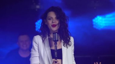 Şık bir vokal kız klasik bir mikrofonda sahnede şarkı söylüyor. Dumanlı ve neon ışıklı karanlık stüdyo. Dinamik neon ışık efektleri. Performans vokali ve müzik grubu. Kapat..