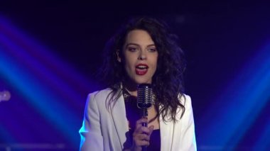 Şık bir vokal kız klasik bir mikrofonda sahnede şarkı söylüyor. Dumanlı ve neon ışıklı karanlık stüdyo. Dinamik neon ışık efektleri. Performans vokali ve müzik grubu. Kapat..