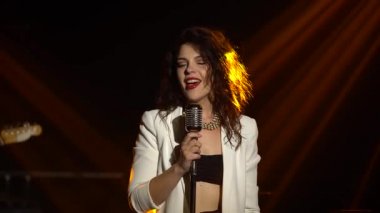 Şık bir vokal kız klasik bir mikrofonda sahnede şarkı söylüyor. Dumanlı ve neon ışıklı karanlık stüdyo. Dinamik neon ışık efektleri. Performans vokali ve müzik grubu. Kapat..