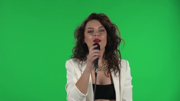 Портрет стильной девушки вокалистки смотрит в камеру и поет. Брюнетка с вьющимися волосами и ярко-красными губами в белой куртке на зеленом экране в студии. — стоковое видео