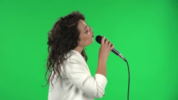 Portret stylowej wokalistki śpiewa i tańczy. Brunetka z kręconymi włosami i jasnoczerwonymi ustami w białej kurtce na zielonym ekranie w studiu. Widok boczny. — Wideo stockowe