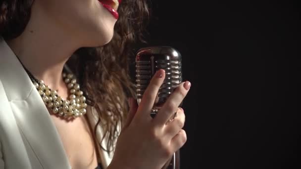 Ένα κομψό κορίτσι τραγουδιστής τραγουδά στη σκηνή σε ένα vintage μικρόφωνο. Σκούρο στούντιο με καπνό και νέον φωτισμό. Λαμπερά κόκκινα χείλη όμορφα σγουρά σκούρα καστανά μαλλιά και καλοφτιαγμένα χέρια κοντά. Δυναμικό νέον — Αρχείο Βίντεο
