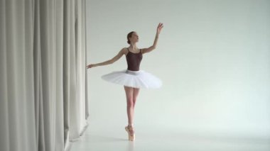 Beyaz balerin etekli balerin dans stüdyosunda pratik yapıyor. Genç kız klasik pas dansı yapıyor ve elleriyle düzgün hareketler yapıyor. Parlak ve ferah bir stüdyoda çekildi..
