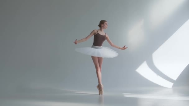 En professionell ballerina i en vit tutu dansar i en stor träningssal. Flickan dansar steg i scenkostymen. Skjuten på en vit bakgrund i den rymliga och starkt upplysta studion. — Stockvideo