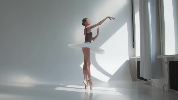 Stüdyoda pencerenin yanında duran genç balerin eğitimi. Beyaz arka planda esnek bir kız parlak ışıkta. — Stok video