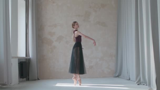 Młoda balerina w czarnej przezroczystej spódniczce w stylu studio na poddaszu stojąca przy oknie. Elastyczna dziewczyna na tle jasnej ściany w jasnym świetle. — Wideo stockowe