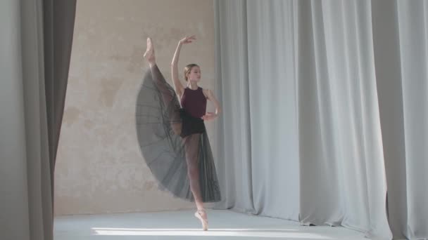 Seorang balerina muda anggun dengan latar belakang tirai cahaya panjang mengangkat kakinya tinggi. Latihan tari bergerak di studio yang luas dan terang benderang. — Stok Video