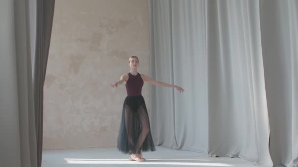 Jeune ballerine gracieuse sur le fond de longs rideaux lumineux effectue un mouvement de pirouette. Répétition danse se déplace dans le studio spacieux et lumineux. — Video
