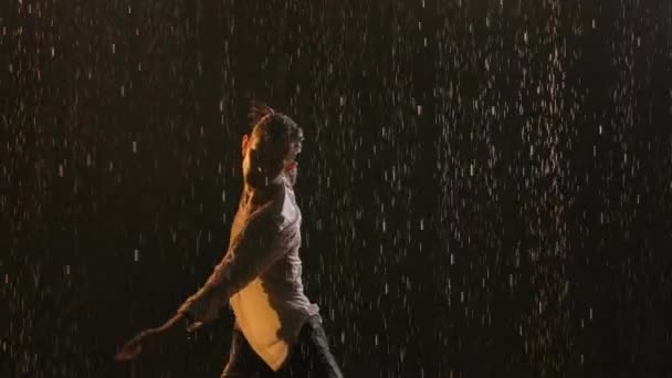 Joven deportista sexy bailando bajo la lluvia en una camisa blanca desabotonada. La ropa húmeda abraza el cuerpo muscular. Las gotas de lluvia brillan en la luz del estudio sobre un fondo negro. De cerca. Movimiento lento. — Vídeos de Stock