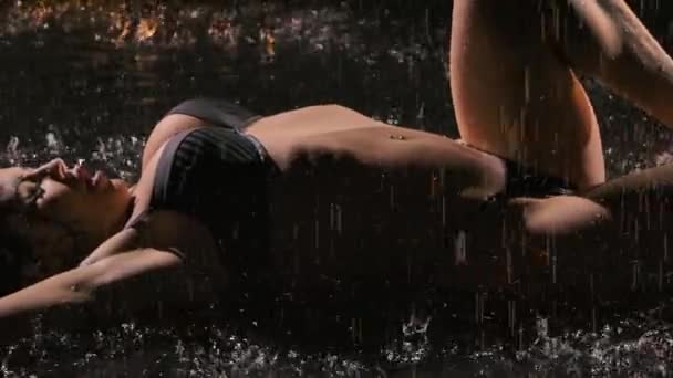 Hermosa morena sexy posando sobre la superficie del agua bajo la lluvia. Cuerpo femenino sexy joven en lencería en gotas de agua que brillan en la luz del estudio. De cerca. Movimiento lento. — Vídeo de stock