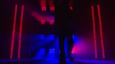 Dans stüdyosunda genç erkek dansçı eğitimi. Dans figürleri yaparken kameraya bakan yakışıklı hip-hop dansçısı. Stüdyoda siyah siluet, duman ve çok renkli neon ışıklı..