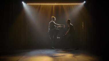 Kaslı bir erkek jimnastikçi asistanının elini iterek darbe yapıyor. Stüdyodaki iki adamın siluetleri sahne ışıklarının arka planına karşı. Yavaş çekim.