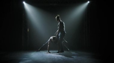 Dengede duran iki jimnastik akrobatik adam. Takım çalışmasında karmaşık numaralar yapan güçlü atletik adamların siluetleri. Siyah bir arka planda sahne ışıkları altında. Yavaş çekim.