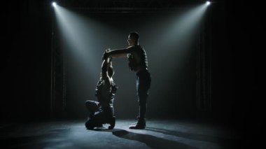 Dengede duran iki jimnastik akrobatik adam. Takım çalışmasında karmaşık numaralar yapan güçlü atletik adamların siluetleri. Siyah bir arka planda sahne ışıkları altında. Yavaş çekim.